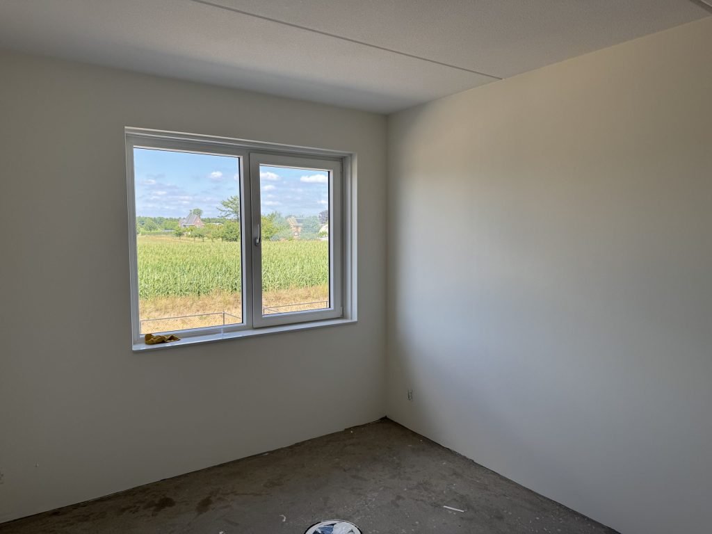 Lege nieuwbouwkamer met strak gesausde witte wanden en een groot raam met uitzicht op een maisveld