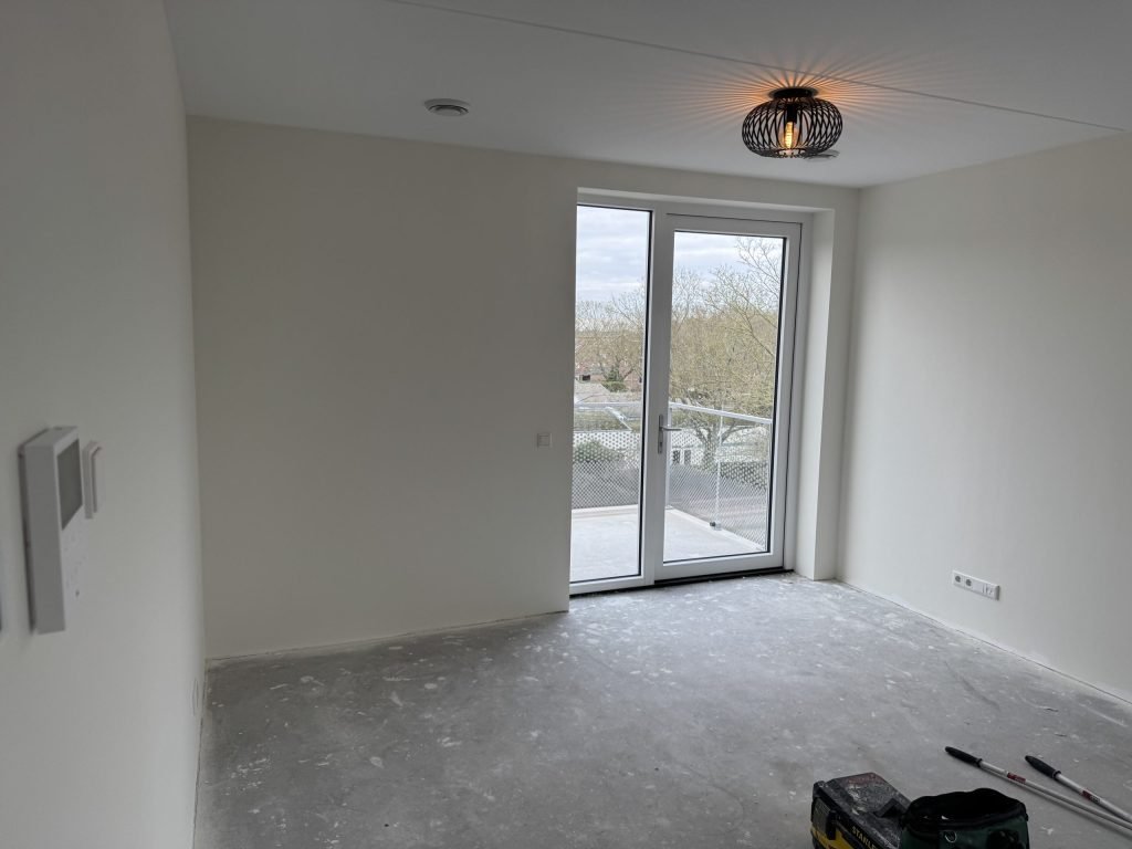Lichte nieuwbouwkamer met strak gesausde wanden, een balkondeur en gereedschap op de betonvloer