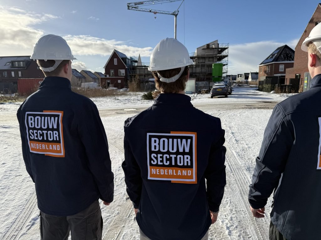 Drie vakmensen van Bouwsector Nederland met witte helm en logo op de jas, op een besneeuwde straat tussen nieuwbouwhuizen